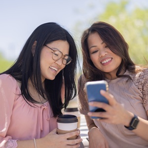 Two women sitting outdoors, smiling and sharing a moment as one shows something on a smartphone.